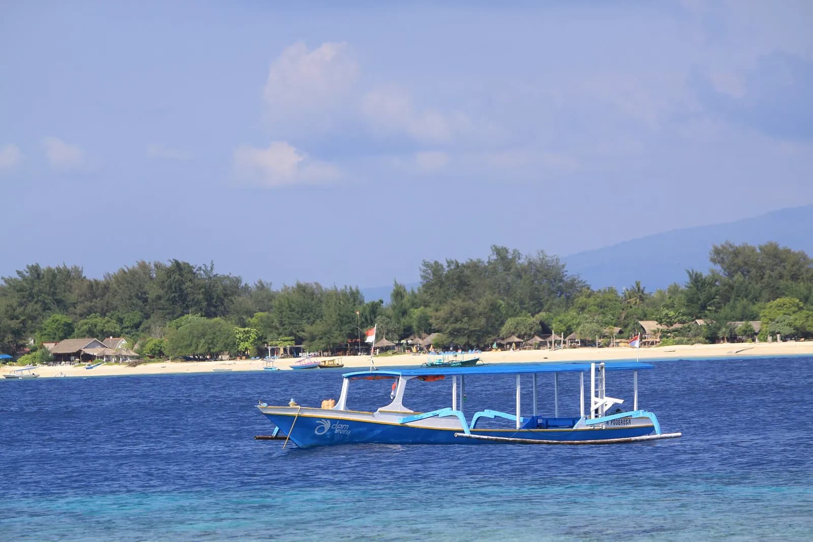Bateau de plongée à balancier ancré devant une plage de sable blanc bordée d'arbres, dans les îles Gili