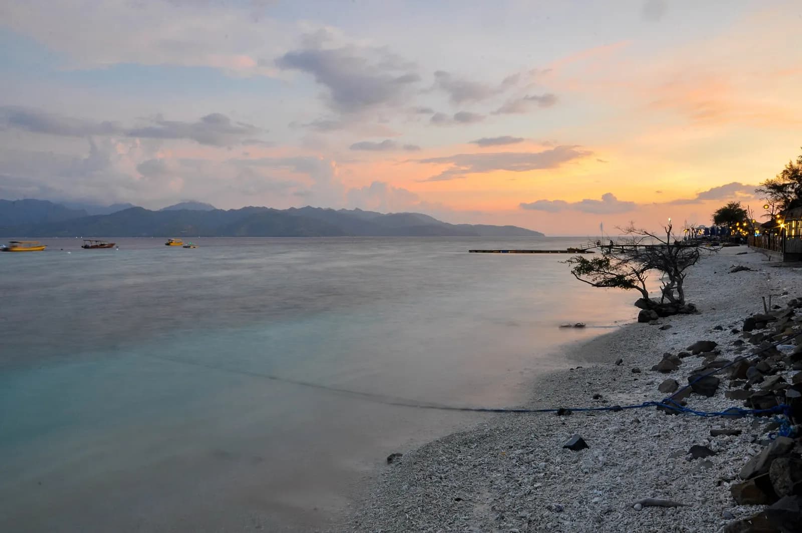 Ciel orangé au coucher du soleil sur une plage de galets et de corail de Gili Trawangan, avec des montagnes à l'horizon
