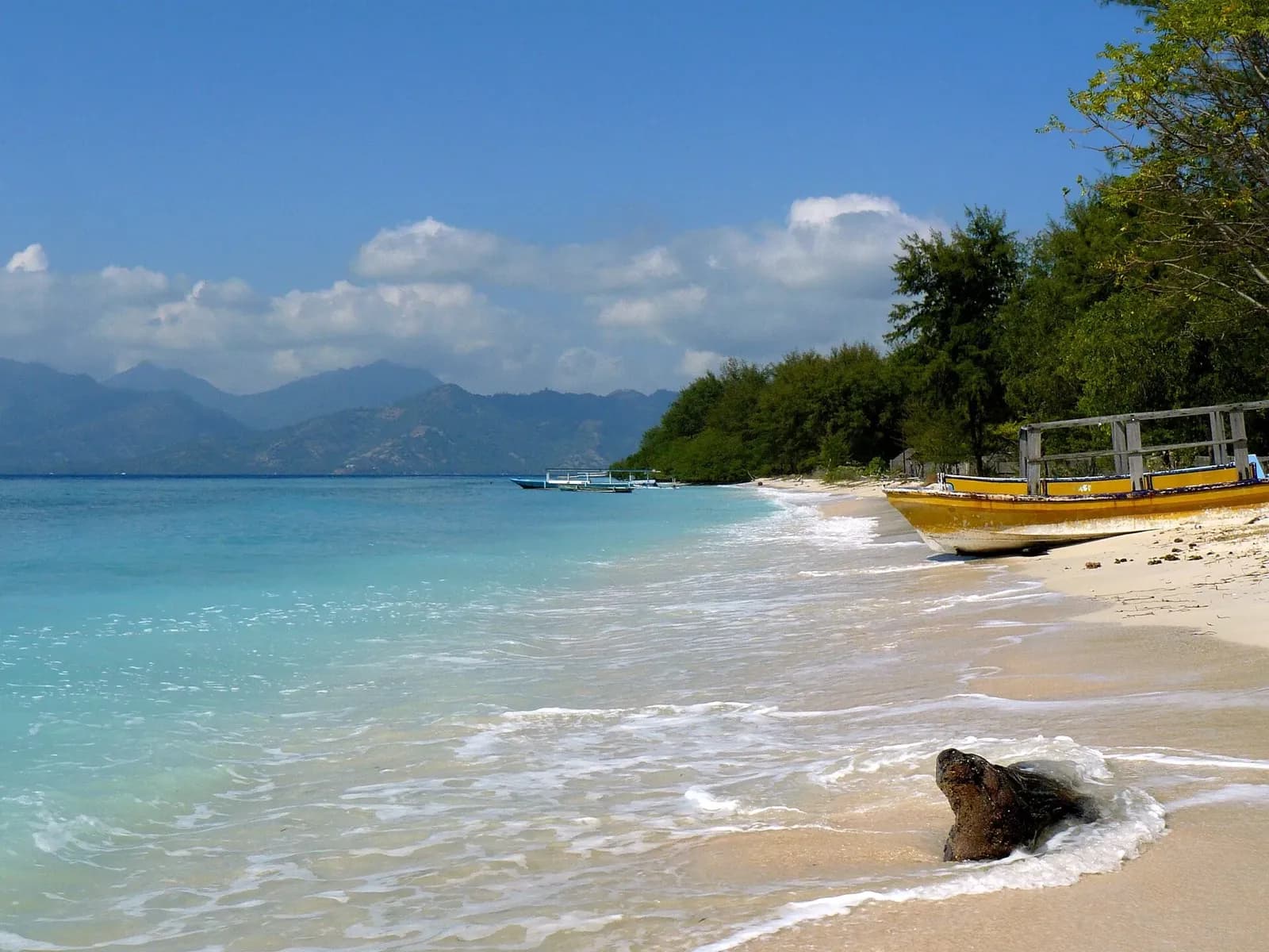 Plage de sable clair et eau turquoise sur Gili Meno, avec une barque échouée et les reliefs de Lombok au fond