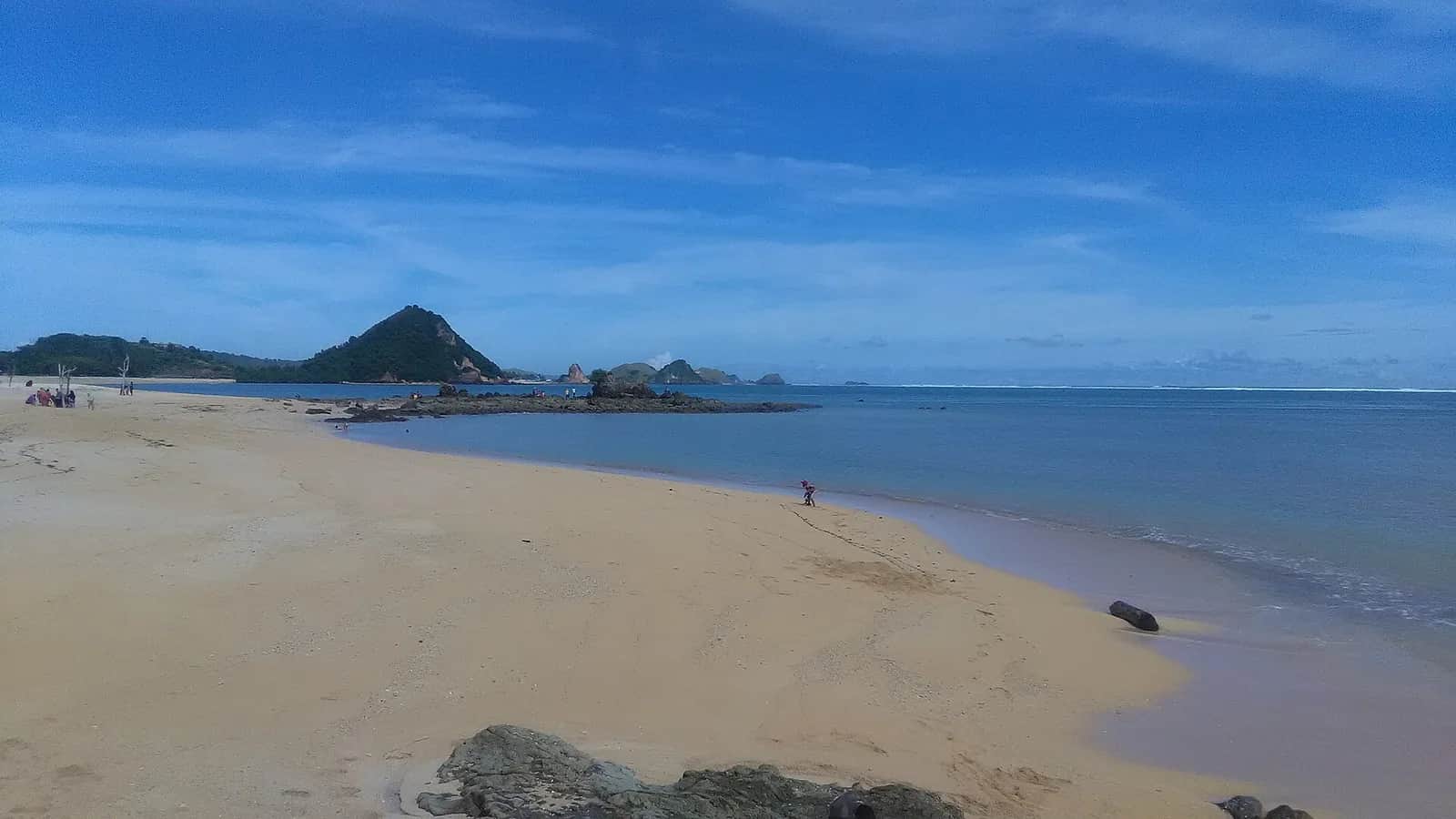 Plage de sable clair de Kuta Lombok sous un ciel bleu, avec les collines de la baie au fond