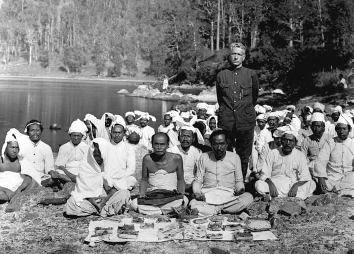Fidèles en turban assis en rang avec leurs offrandes au bord du lac Segara Anak, au Rinjani, lors de la fête annuelle photographiée vers 1925, avec un homme debout au centre