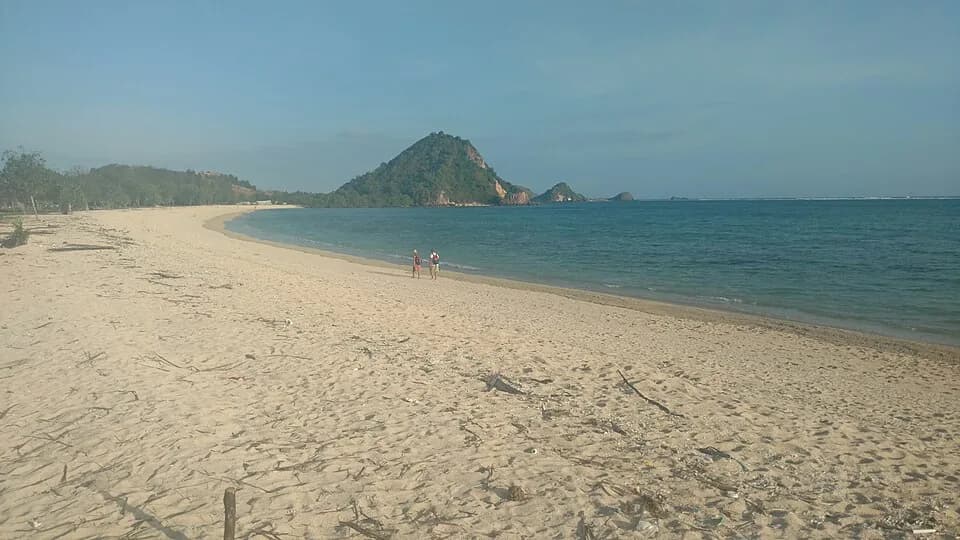 Longue plage de sable clair presque déserte devant un promontoire boisé, sur la côte de Lombok